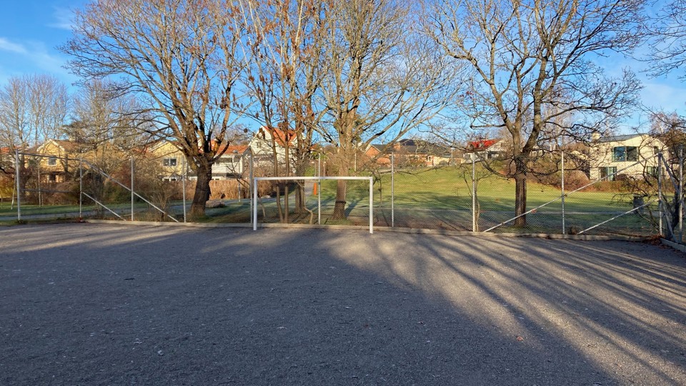 En grusplan med ett fotbollsmål. Bakom planen syns träd och hus.