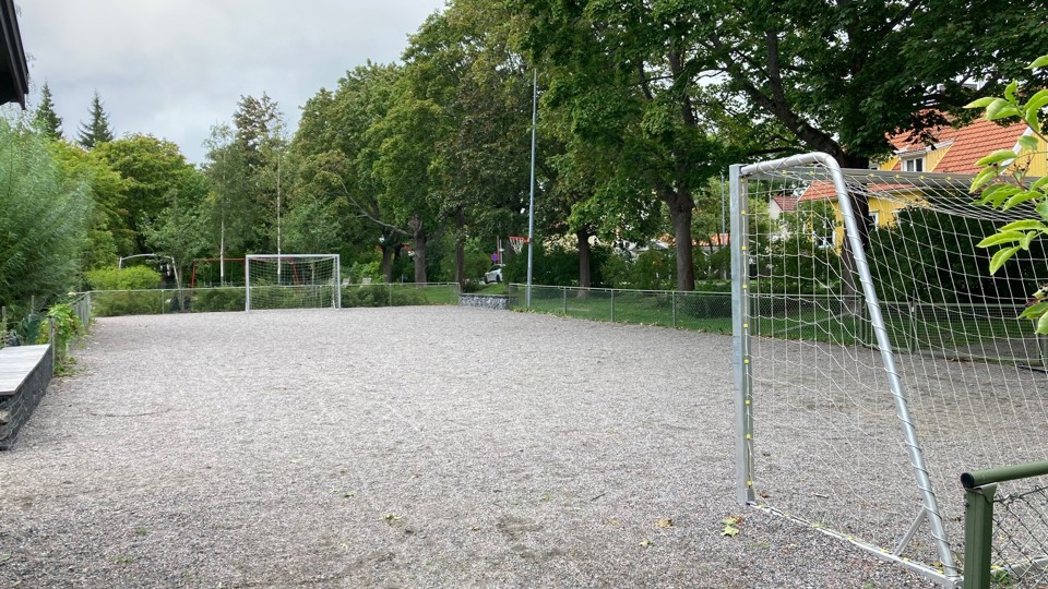 Grusbelagd bollplan med fotbollsmål, omgiven av träd och ett staket. I bakgrunden syns ett gult hus.