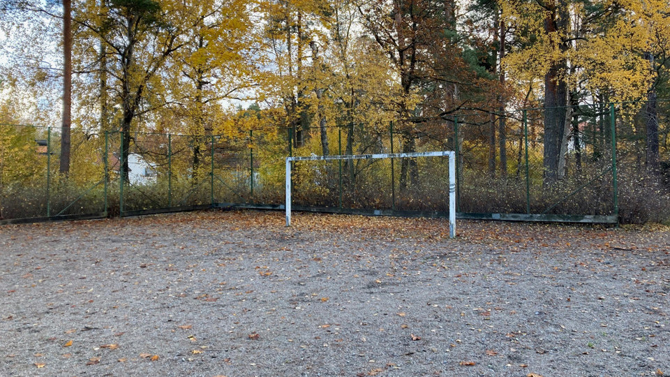 Grusplan omgärdad av högt stängsel. Ett fotbollsmål.