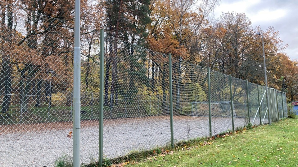 Inhägnad grusplan med fotbollsmål och träd i höstfärger bakom.