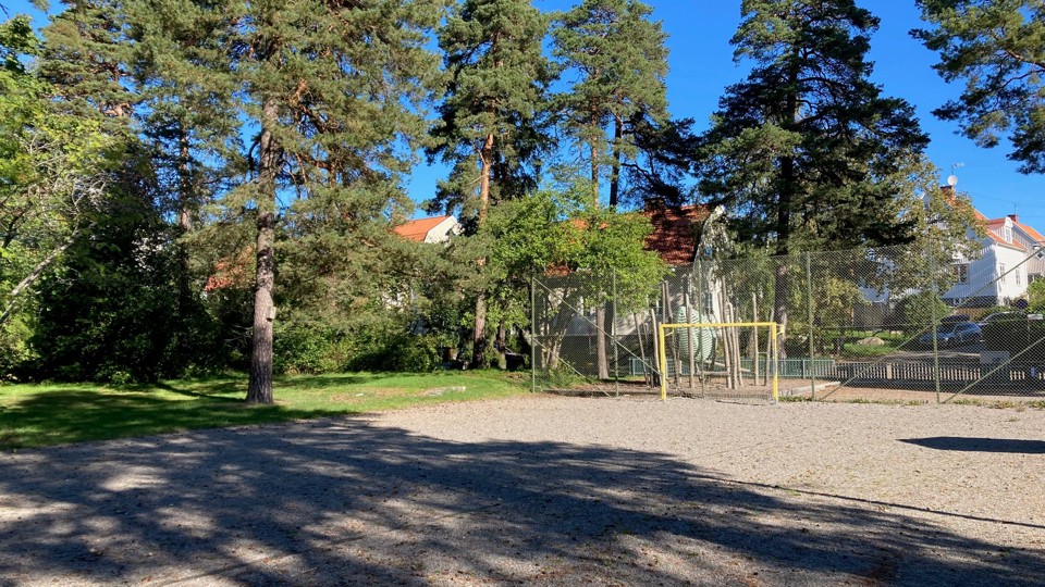 En grusplan med fotbollsmål, skuggad av höga träd och inhägnad med ett stängsel, med hus i bakgrunden.