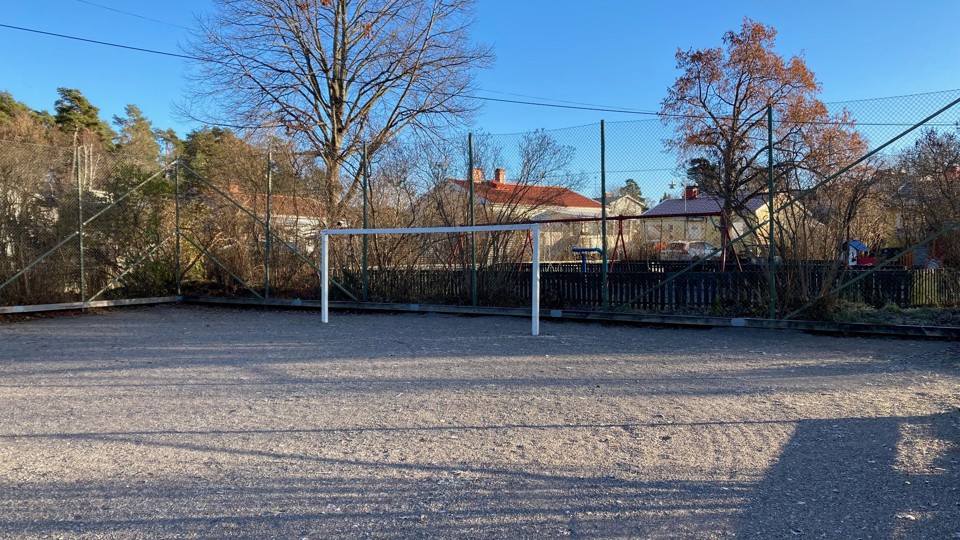 Grusbelagd bollplan med fotbollsmål, omgärdad av stängsel. I bakgrunden en lekpark och flera hus.