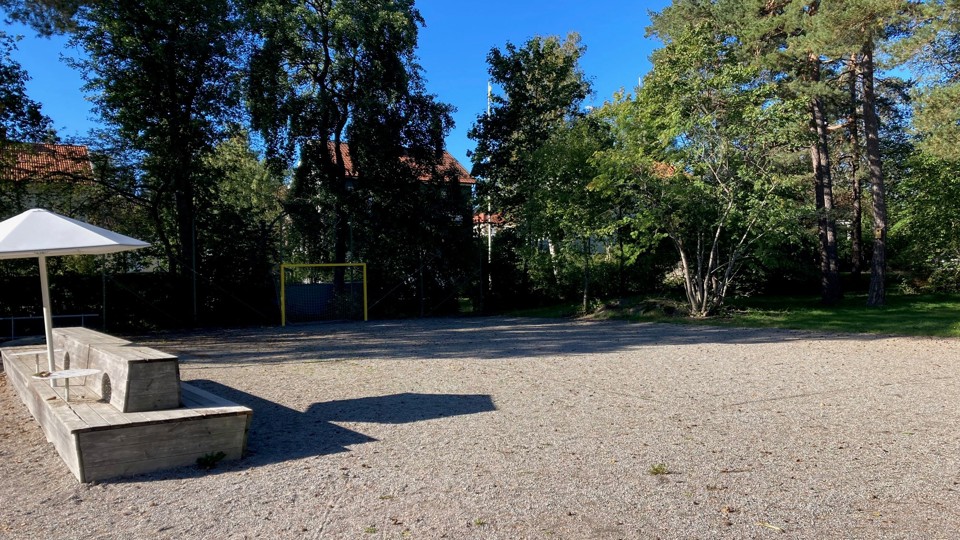 En grusad fotbollsplan med träd och ett fotbollsmål i bakgrunden, med en skuggig sittplats.