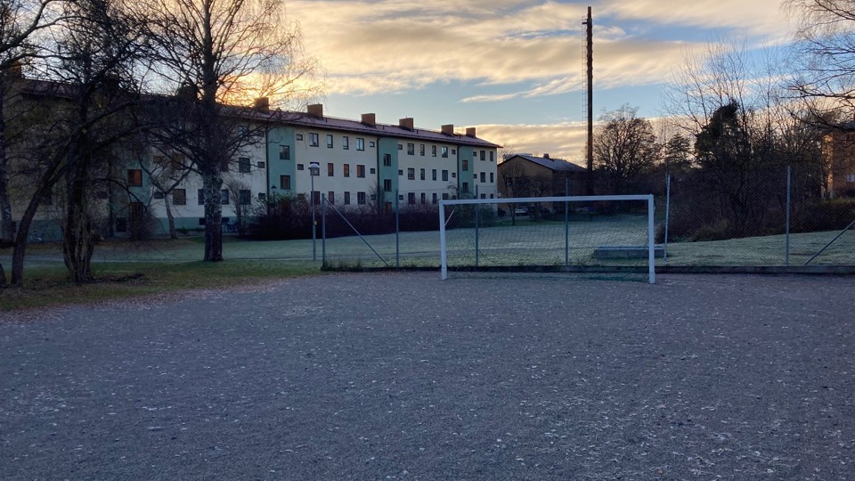 En grusplan med målstolpar och ett staket. I bakgrunden syns flerbostadshus och träd, med soluppgång på himlen.