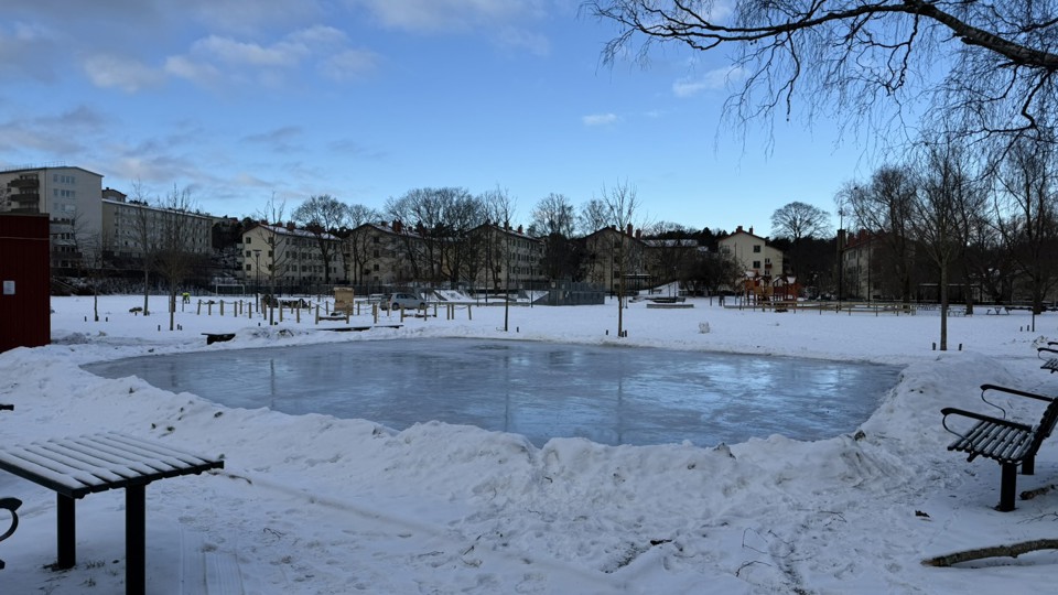 I förgrunden etta par träbänkar. En skridskois omgiven av snö och träd som står glest. Vid kanten på av isen finns träbänkar. I bakgrunden flera flerbostadshus. Foto.