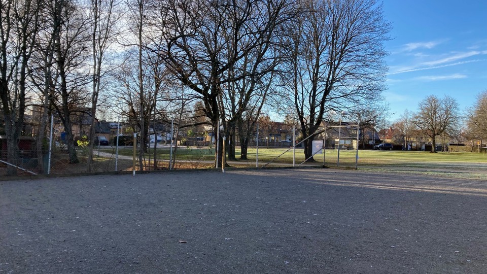 En grusplan omgiven av träd och stängsel. I bakgrunden finns flera hus.