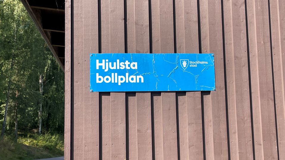 En blå skylt som det står Hjulsta bollplan på sitter på väggen till ett brunt förråd.