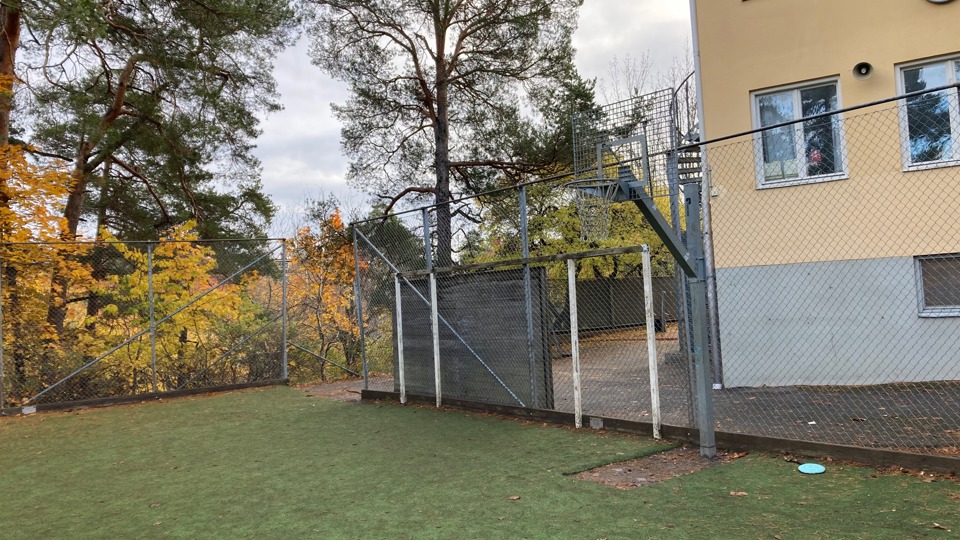 Bollplan med basketkorg och ett mindre fotbollsmål intill skolbyggnad, omgiven av stängsel.