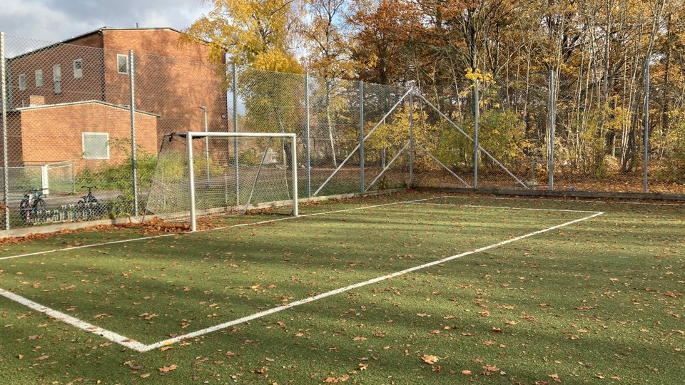 En målbur på en inhägnad bollplan med konstgräs, träd och en tegelbyggnad bakom.