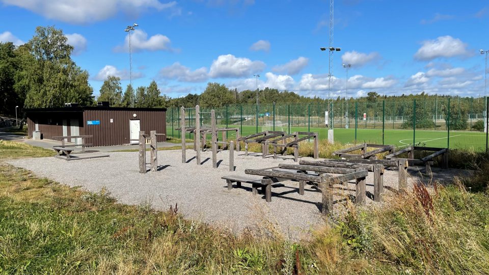 Gymredskap i trä som står på ett grusunderlag invid en fotbollsplan.