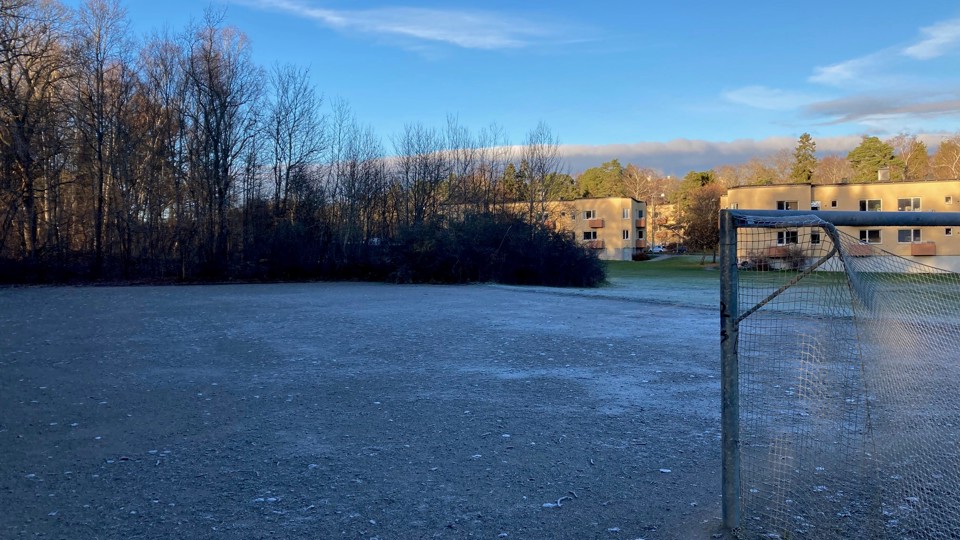 Frosttäckt grusplan med fotbollsmål och bostadshus i bakgrunden.
