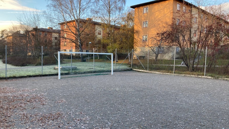 En grusplan med ett fotbollsmål och träd runtom, med en byggnad i bakgrunden.