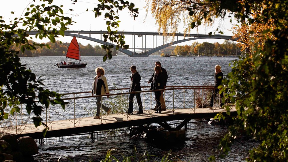 Personer som promenerar på en liten gångbro vid vattnet. I bakgrunden syns Västerbron.