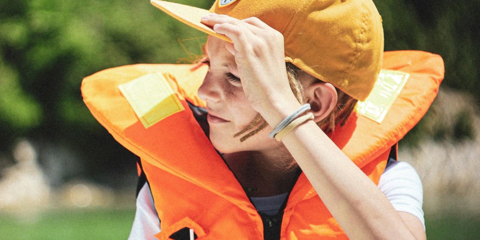 En ung person med en orange räddningsflytväst och en orange keps vid en sjö.