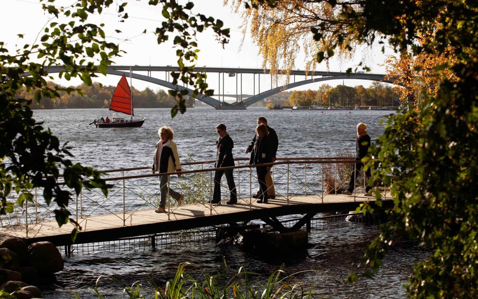 Personer som promenerar på en liten gångbro vid vattnet. I bakgrunden syns Västerbron.