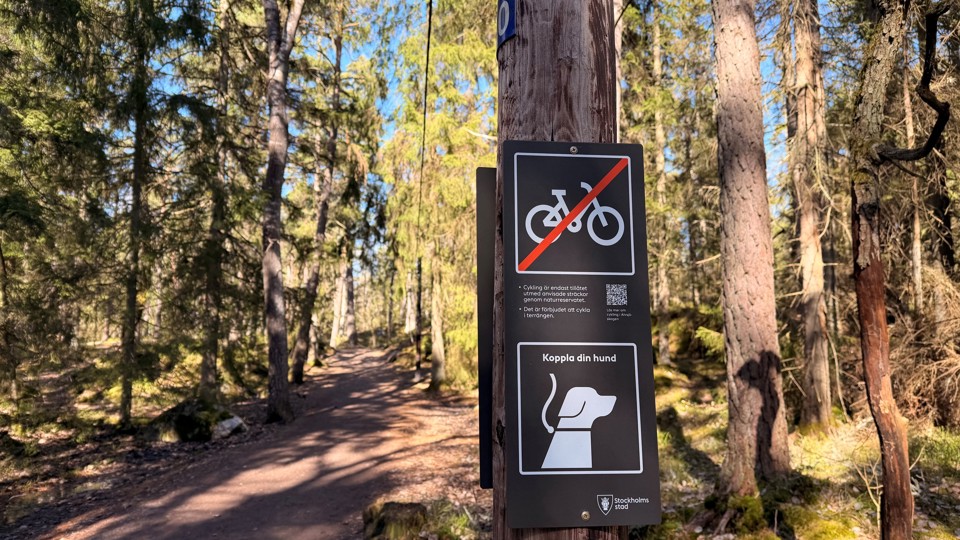 En grusväg leder fram genom skogen. På en stolpe sitter en skylt som visar en kopplad hund och en överkryssad cykel.