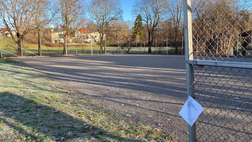 En grusplan med ett fotbollsmål. Runt planen finns stängsel, träd och några hus i bakgrunden.