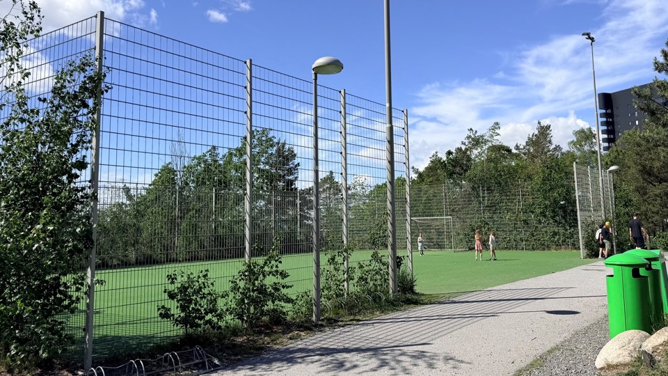En fotbollsplan med högt stängsel, barn och vuxna på planen samt en gångväg i en grön parkmiljö under blå himmel.