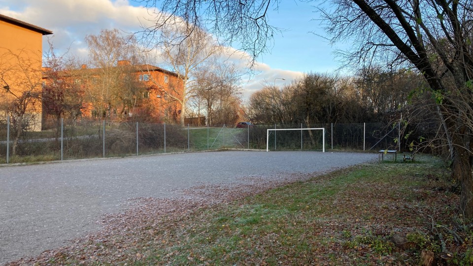 En grusplan med ett fotbollsmål, omgiven av ett stängsel och träd, med en byggnad i bakgrunden.