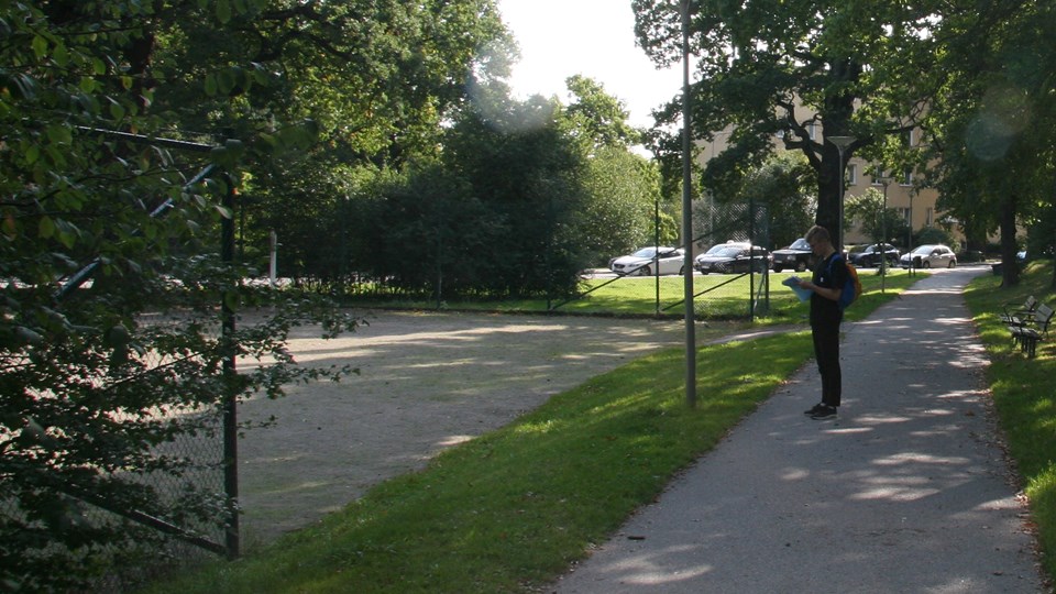 En ungman står på parkväg intill en grusplan.