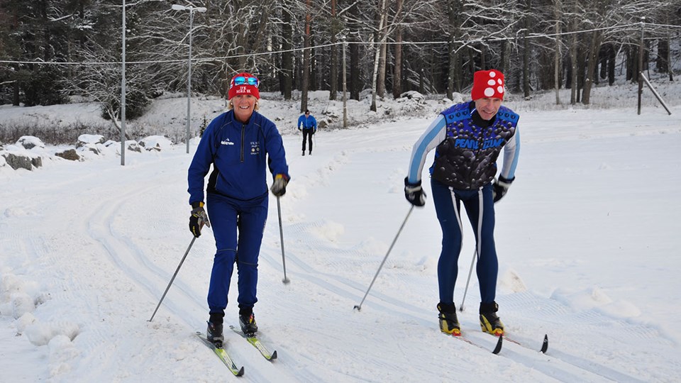 En man och en kvinna i blå kläder och röda mössor åker längdskidor i ett skidspår. Personerna ser glada ut.
