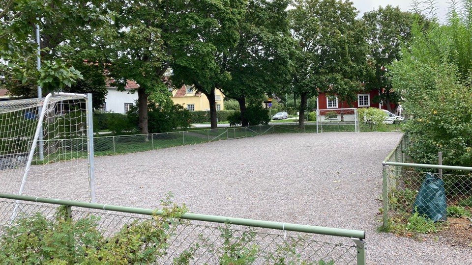 Grusbelagd bollplan med fotbollsmål, omgiven av träd och ett staket. I bakgrunden syns ett rött hus.