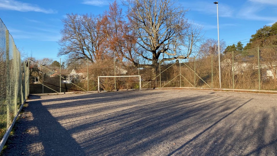 Grusbelagd bollplan med fotbollsmål, omgärdad av stängsel. I bakgrunden syns flera hus.