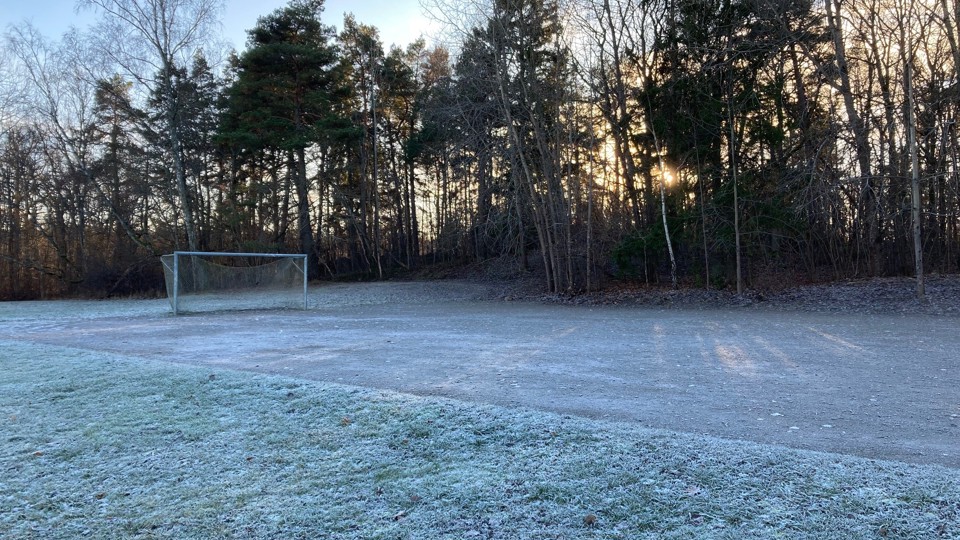 Frosttäckt grusplan med fotbollsmål, omgiven av träd i solljus.