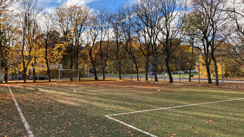 En tom bollplan med konstgräs och höstlöv, omgärdad av stängsel och träd med gula blad.