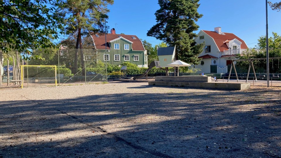 En grusad lekplats med fotbollsmål, gungor och en sittplats under ett tak, omgiven av träd och hus i bakgrunden.